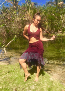 A woman wears a soft cotton lycra two tone skirt that is red and grey with mandala print. The skirt is adjustable in length with sinch options on 4 sides. She is standing in front of palm trees on a beach in Hawaii.